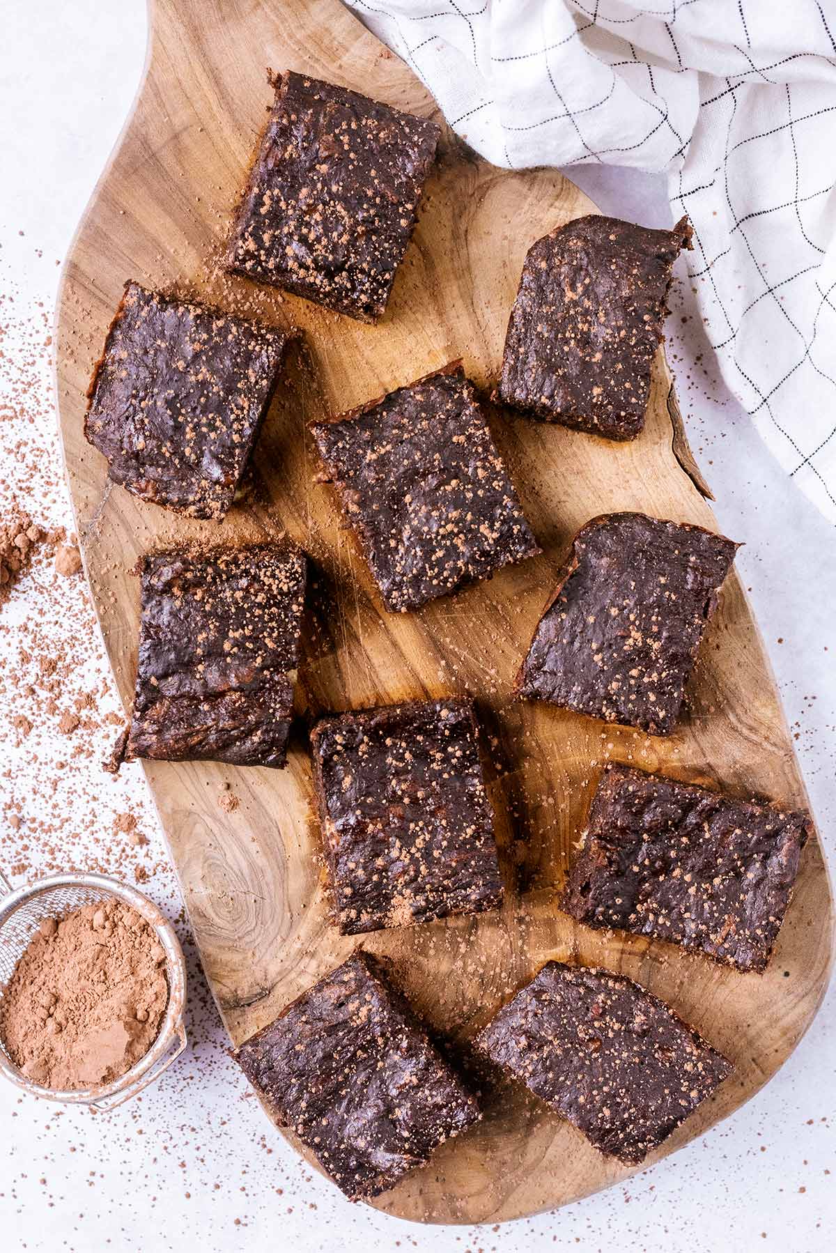 Ten brownies on a wooden serving board next to a small sieve of cocoa powder.