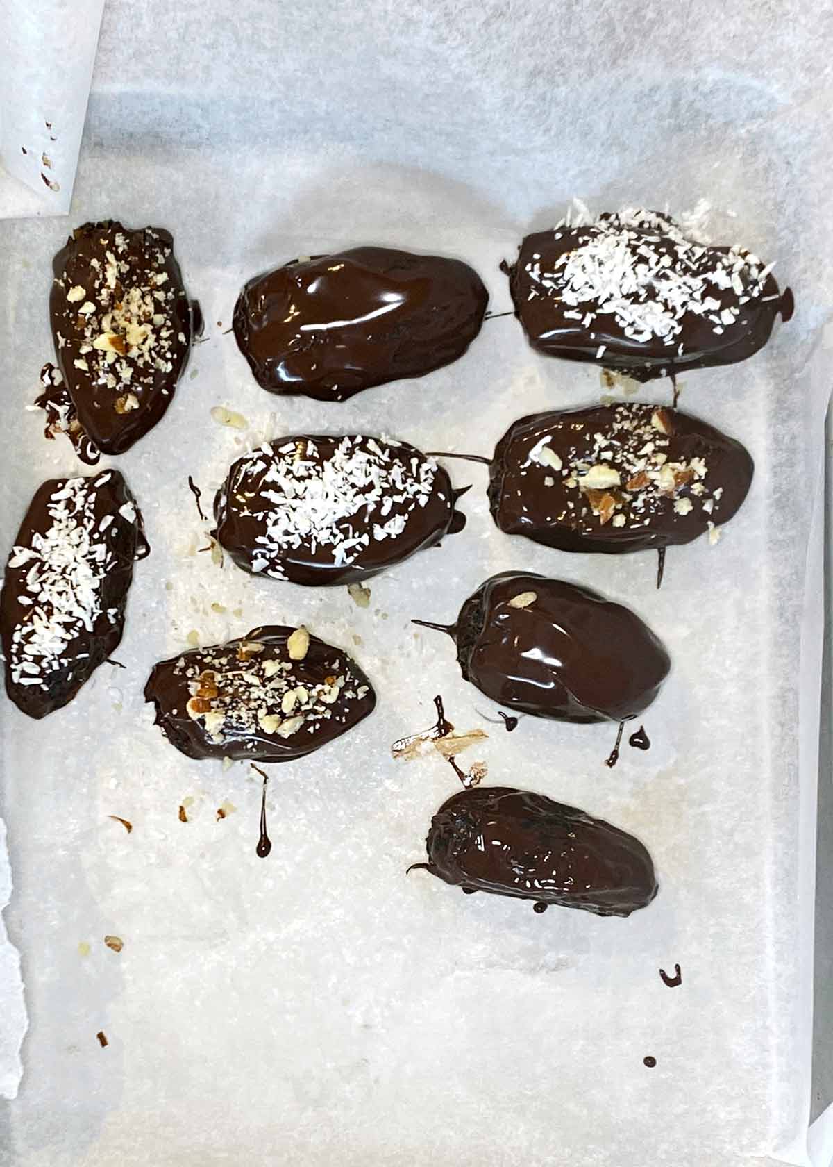 Chocolate covered dates on a lined tray topped with crushed nuts and desiccated coconut.