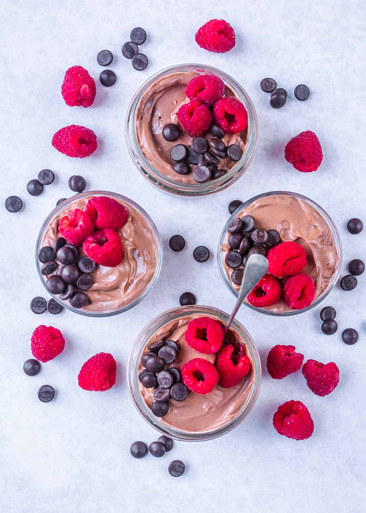 Four pots of chocolate flavored yogurt surrounded by raspberries and chocolate chips.