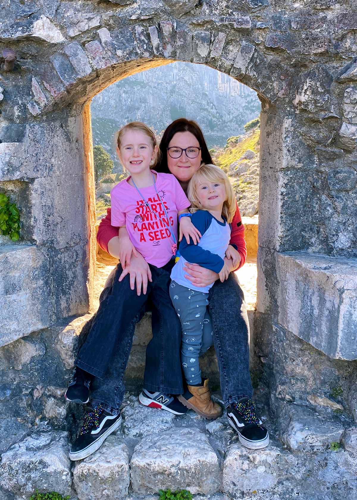 A woman, a female child and a male child sat in a stone window surround.